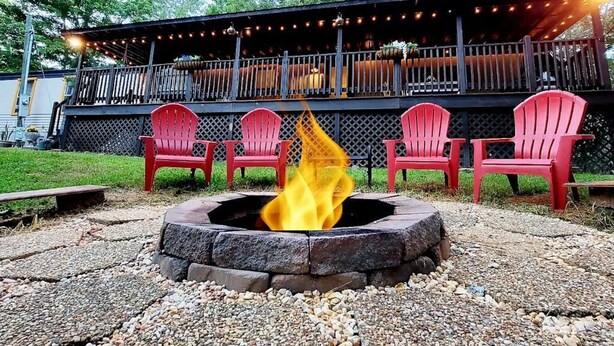 A fire pit with red chairs arranged around it, a covered deck with string lights, and a building in the background. - Inn the Woods - cozy place in the Smokies (Sevierville)