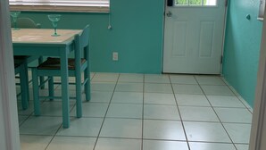 Een kamer met teal muren, een witte deur en een raam met zonwering. Er staat een tafel met stoelen en een decoratief bord op de muur.
