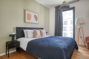 Un dormitorio con cama, mesita de noche, lámpara y lámpara de pie, y una ventana con cortinas.
