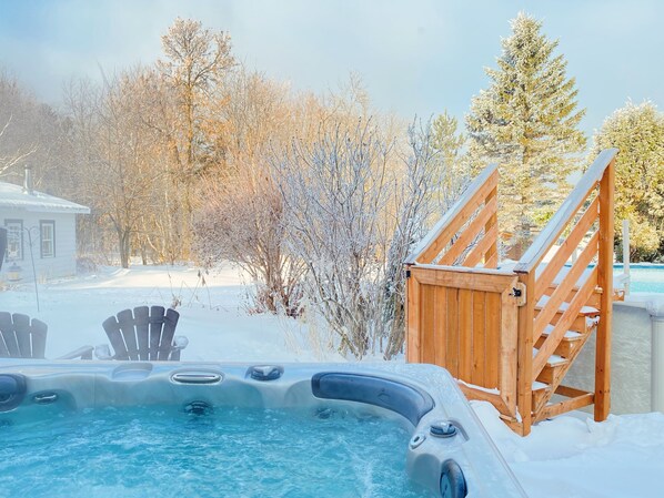 A hot tub with a wooden deck and stairs leading up to it, surrounded by snow-covered trees and a house in the background.