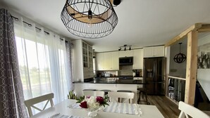 A modern kitchen with white cabinets, a stainless steel refrigerator, and a dining area with a striped tablecloth and white chairs. - Cottage - The Retro   (Saint-Michel-de-Bellechasse)