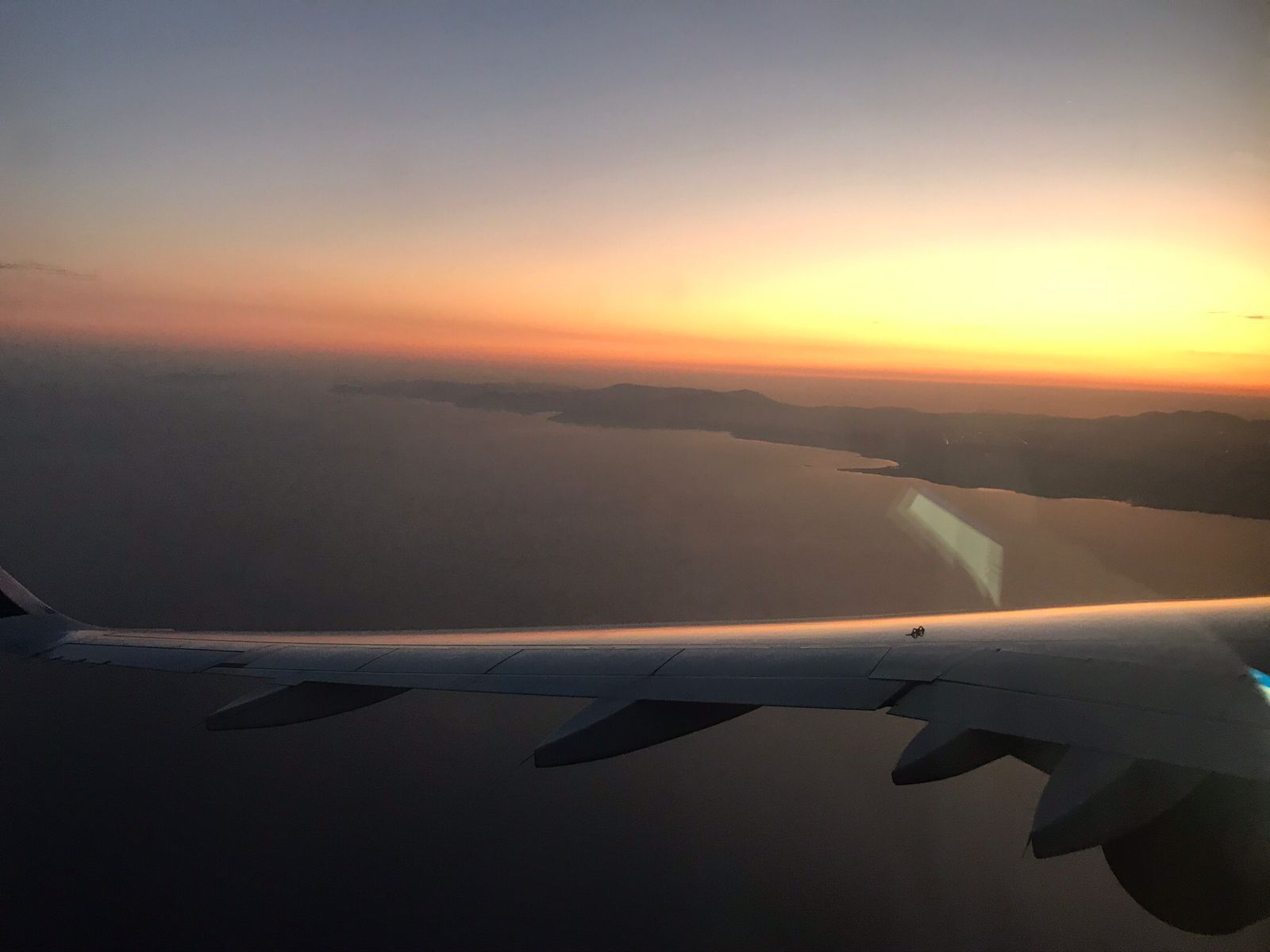 En flygplansvinge med utsikt över en solnedgång över ett vattenområde och ett avlägset landområde.