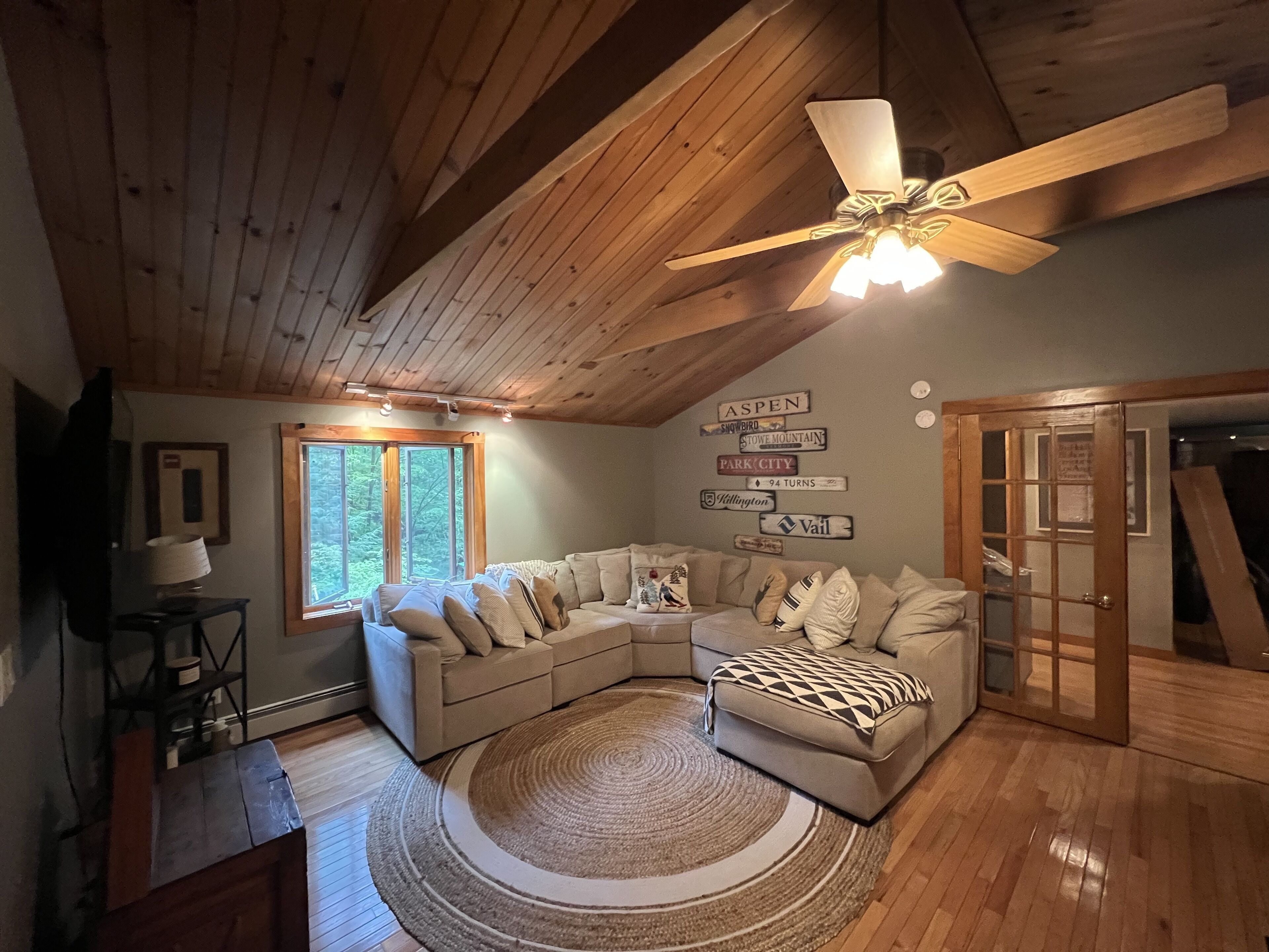 Ein gemütliches Wohnzimmer mit Holzboden, Deckenventilator, großem Fenster, Eckcouch, Couchtisch und Teppich.