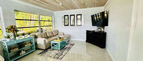 A modern living room with a sofa, coffee table, and TV. There are large windows with a view of greenery outside.