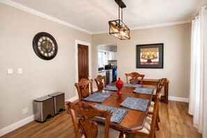 A dining room with a wooden table, chairs, a clock, a pendant light, and a kitchen in the background.