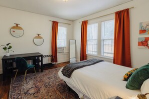 Un dormitorio con cama, escritorio, silla, espejo y ventana con cortinas.