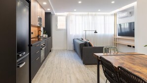 Une cuisine moderne avec des meubles de couleur sombre, un plan de travail en bois et un four encastré. On y trouve un coin repas avec une table et des chaises en bois, et un canapé gris à l’arrière-plan.