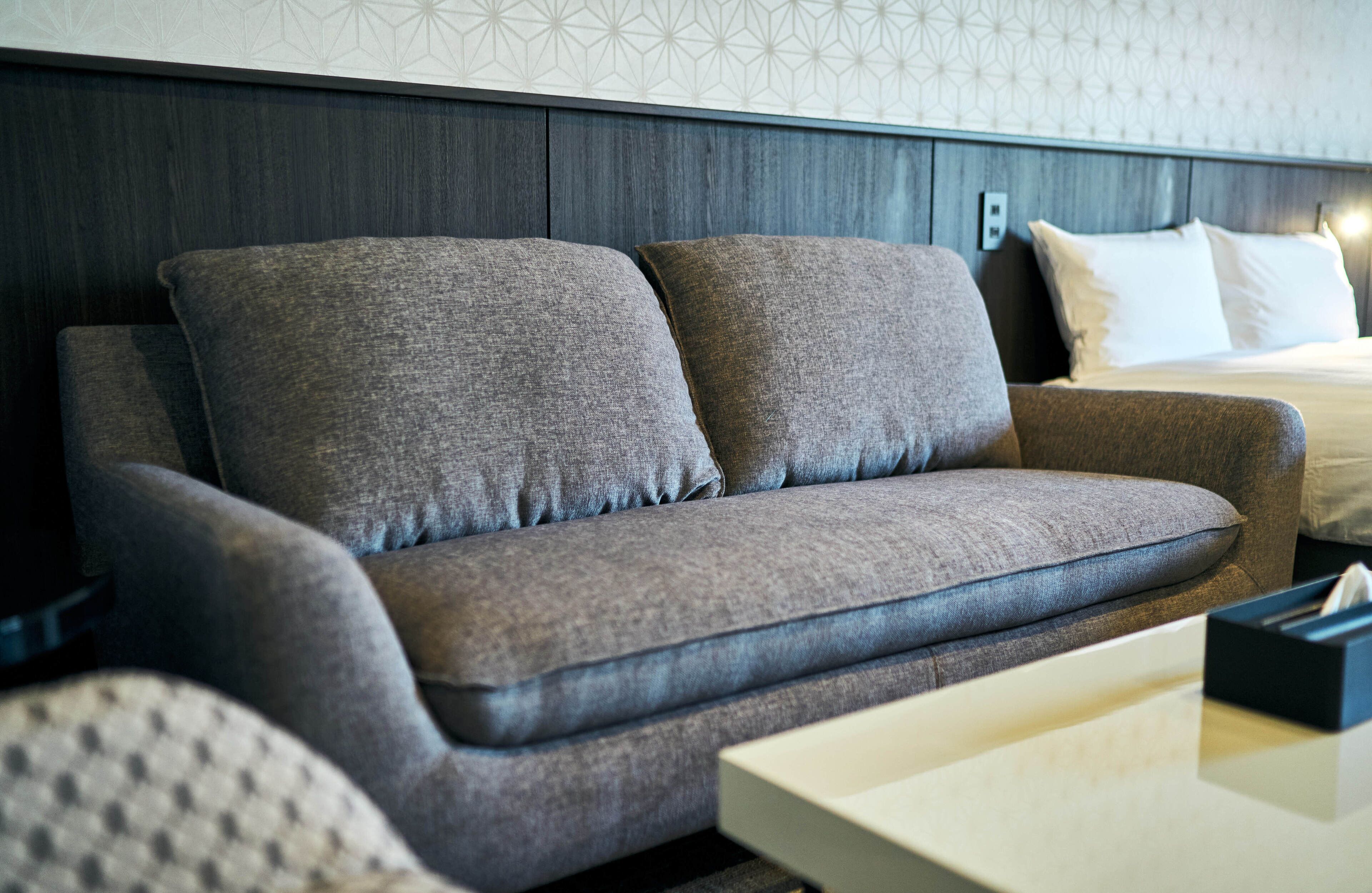 Een moderne hotelkamer met een grijze bank, een witte nachtkastje en een opgemaakt bed met witte lakens.