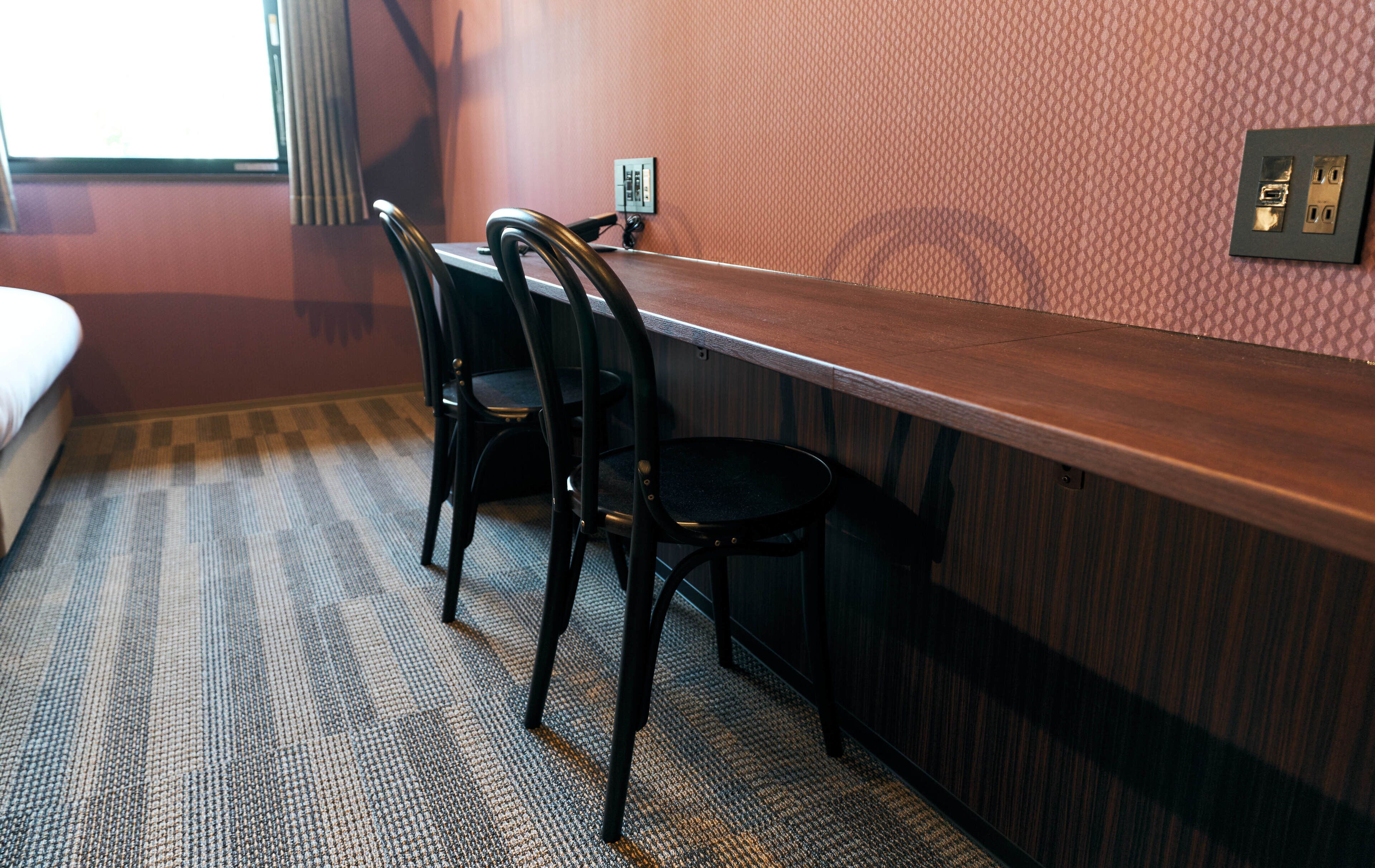 A hotel room with a wooden desk, two black chairs, a bed, and a textured carpet.