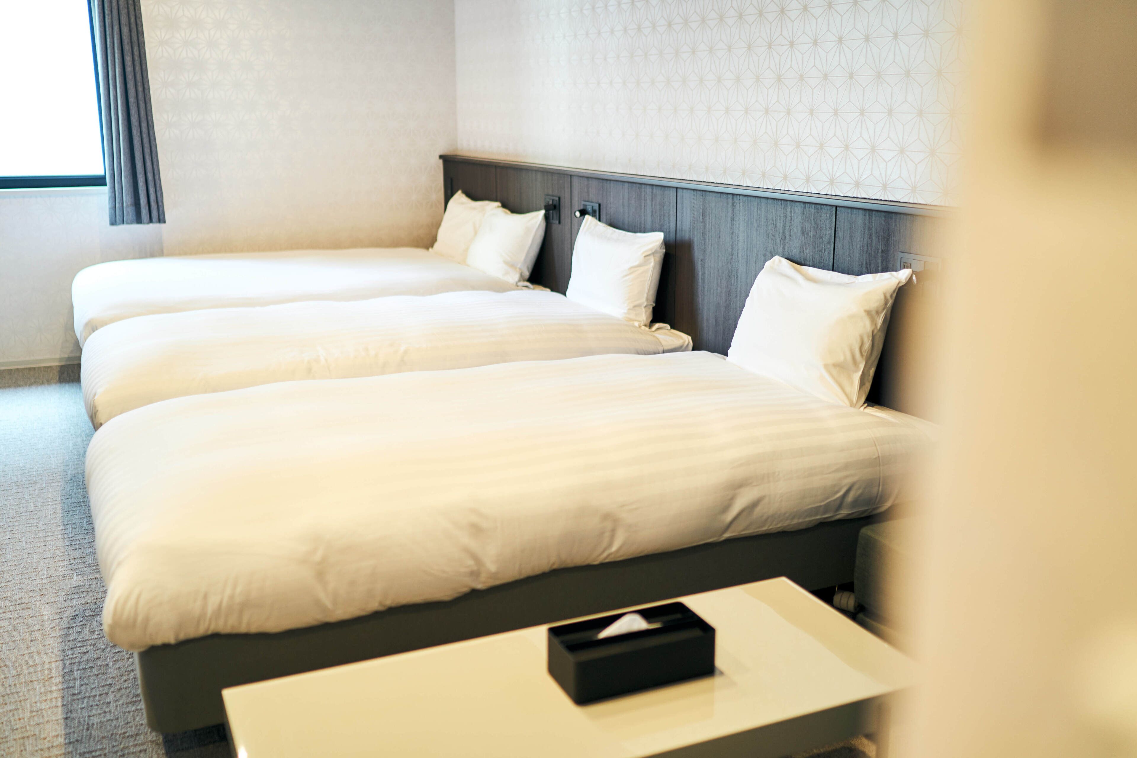 Ein Hotelzimmer mit drei Betten, einem Nachttisch mit Taschentuchbox und einer Wand mit Muster.