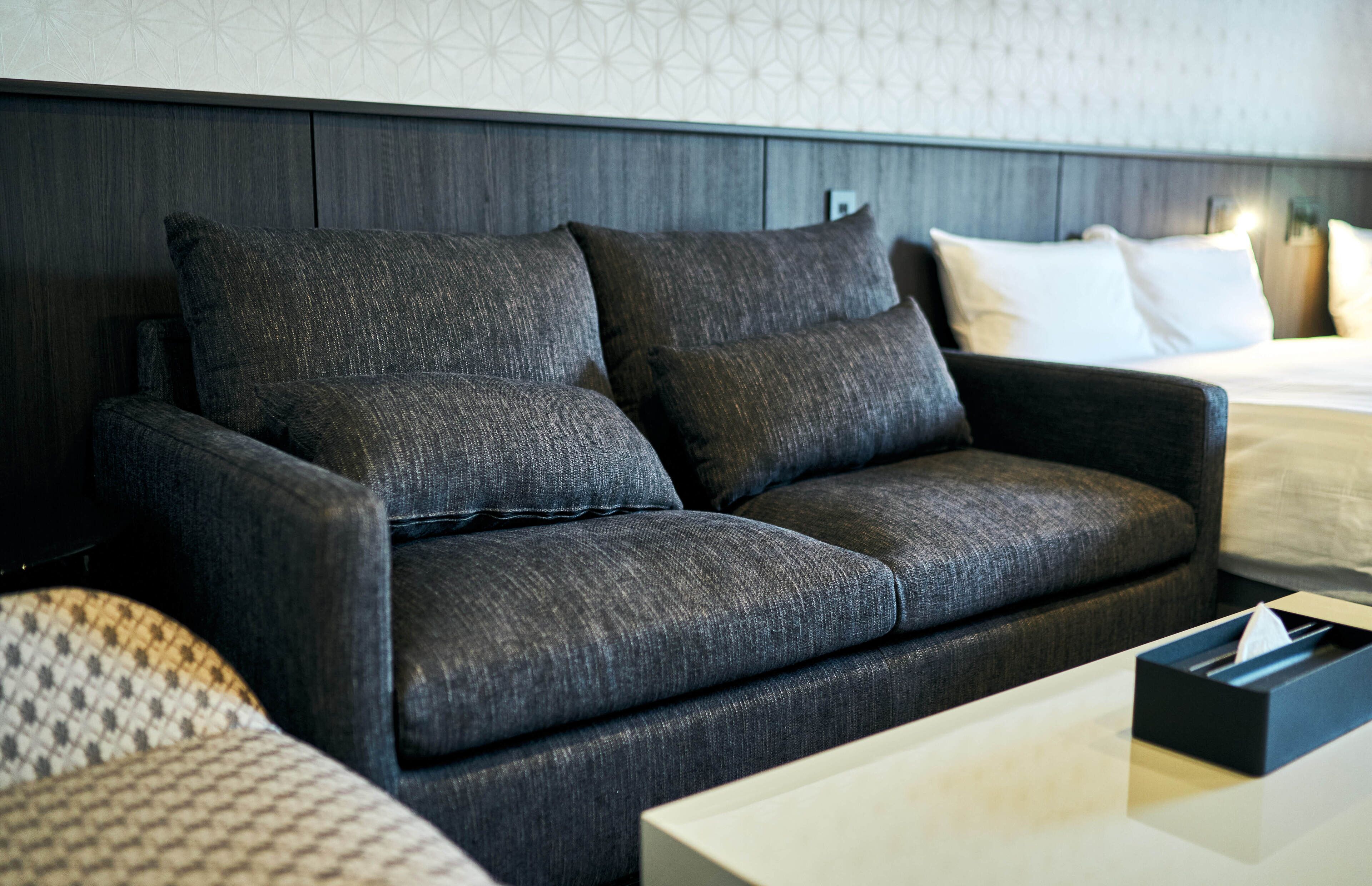 Una habitación de hotel moderna con un sofá gris oscuro, una cama blanca y un cojín estampado.