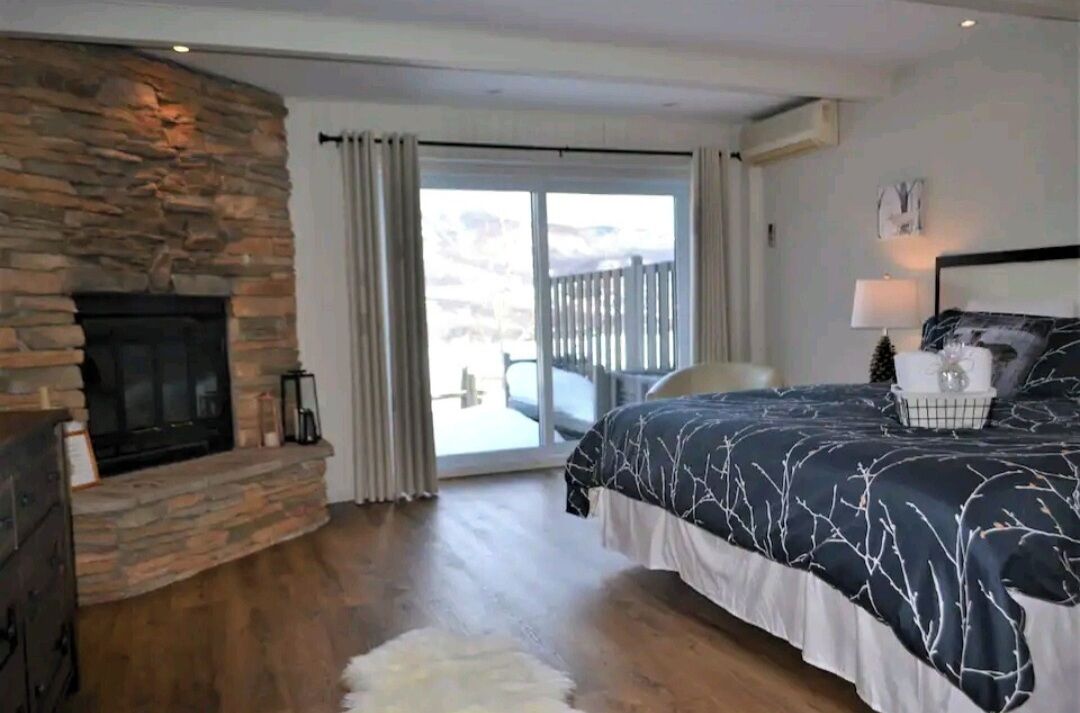 Un dormitorio con chimenea de piedra, una cama con cubrecama estampado con ramas de árbol, una alfombra blanca y mullida, y vistas a un balcón con barandilla de madera.