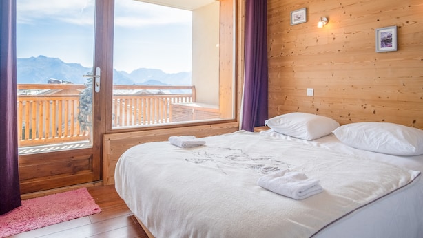 A bedroom with a wooden floor, a bed with white linens, a large window with a view of mountains, and a wooden ceiling. - Appartement du Poutat - Welkeys (Huez)