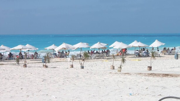 Een strand met witte parasols en plantenbakken, waar mensen in stoelen schaduw zoeken.