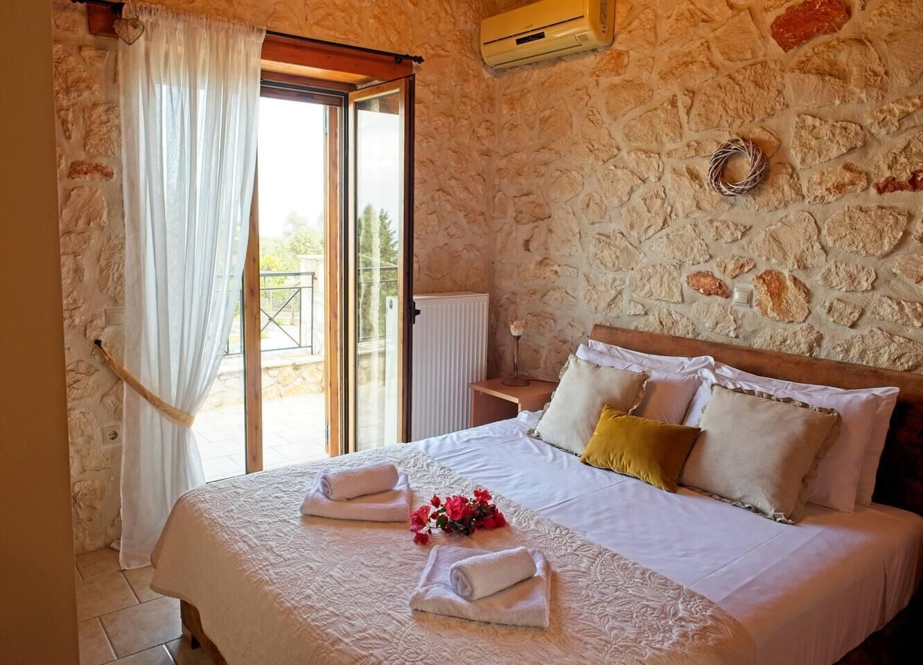 A bedroom with a stone wall, a bed with white linens and pillows, a window with curtains, and a small table with a vase.