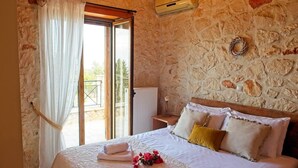 Un dormitorio con pared de piedra, una cama con sábanas y almohadas blancas, una ventana con cortinas y una mesita con un jarrón.