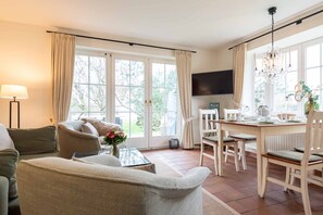 Una habitación luminosa con un sofá, una mesa de comedor con sillas, un televisor y ventanas grandes con cortinas.