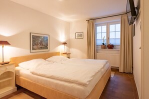 Ein Hotelzimmer mit einem großen Bett, zwei Nachttischen, einem fenster mit Vorhängen, einer Heizung und einem an der Wand montierten Fernseher.