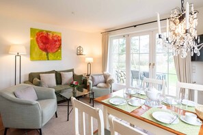 Ein Wohnzimmer mit einer Couch, Sesseln, einem Couchtisch und einem Esstisch, gedeckt für vier Personen. An der Wand hängt ein Gemälde, eine Kristallleuchter und ein Blick durch das Fenster.