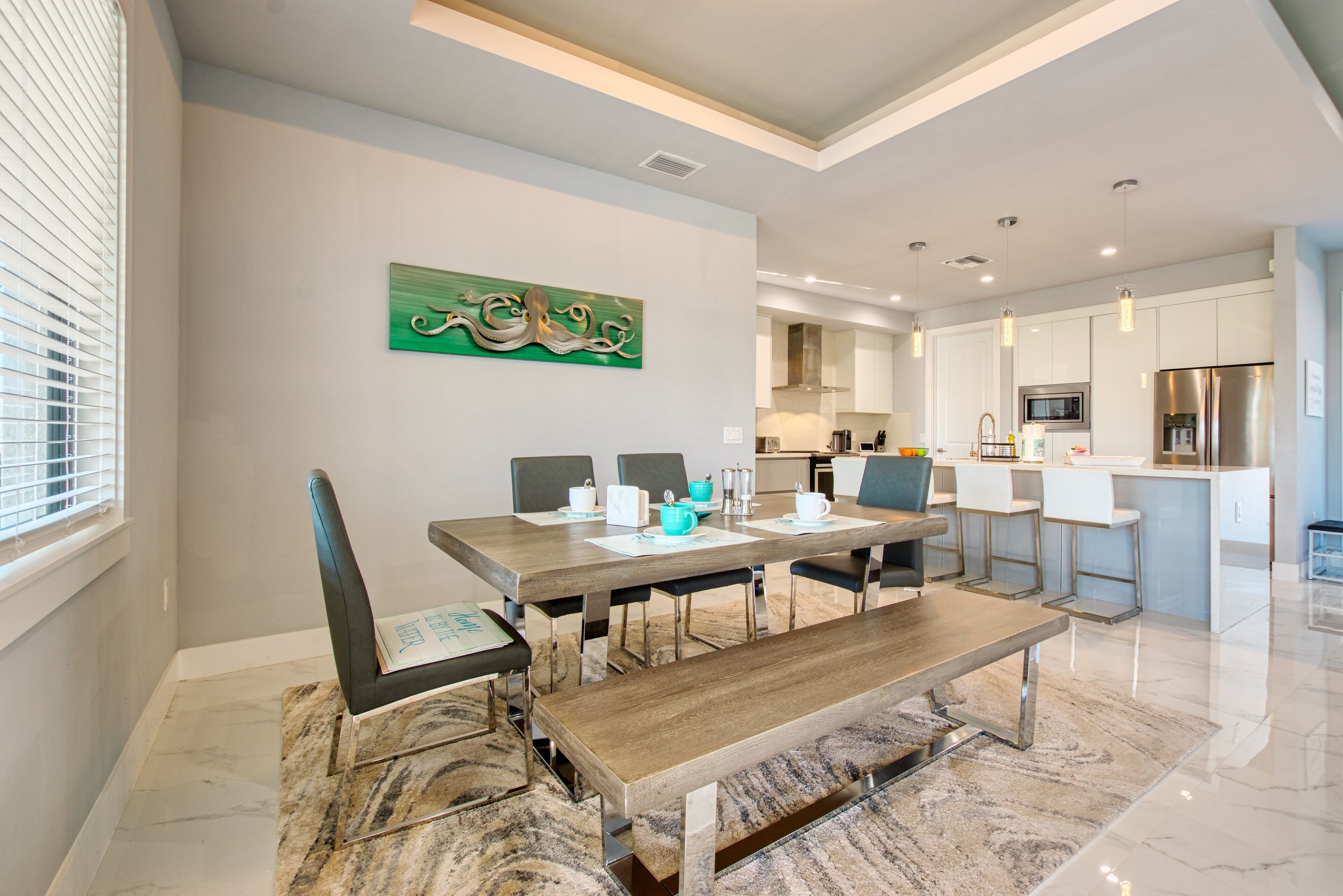 A modern dining area with a wooden table, chairs, and a bench. There is a kitchen in the background with white cabinets and stainless steel appliances.