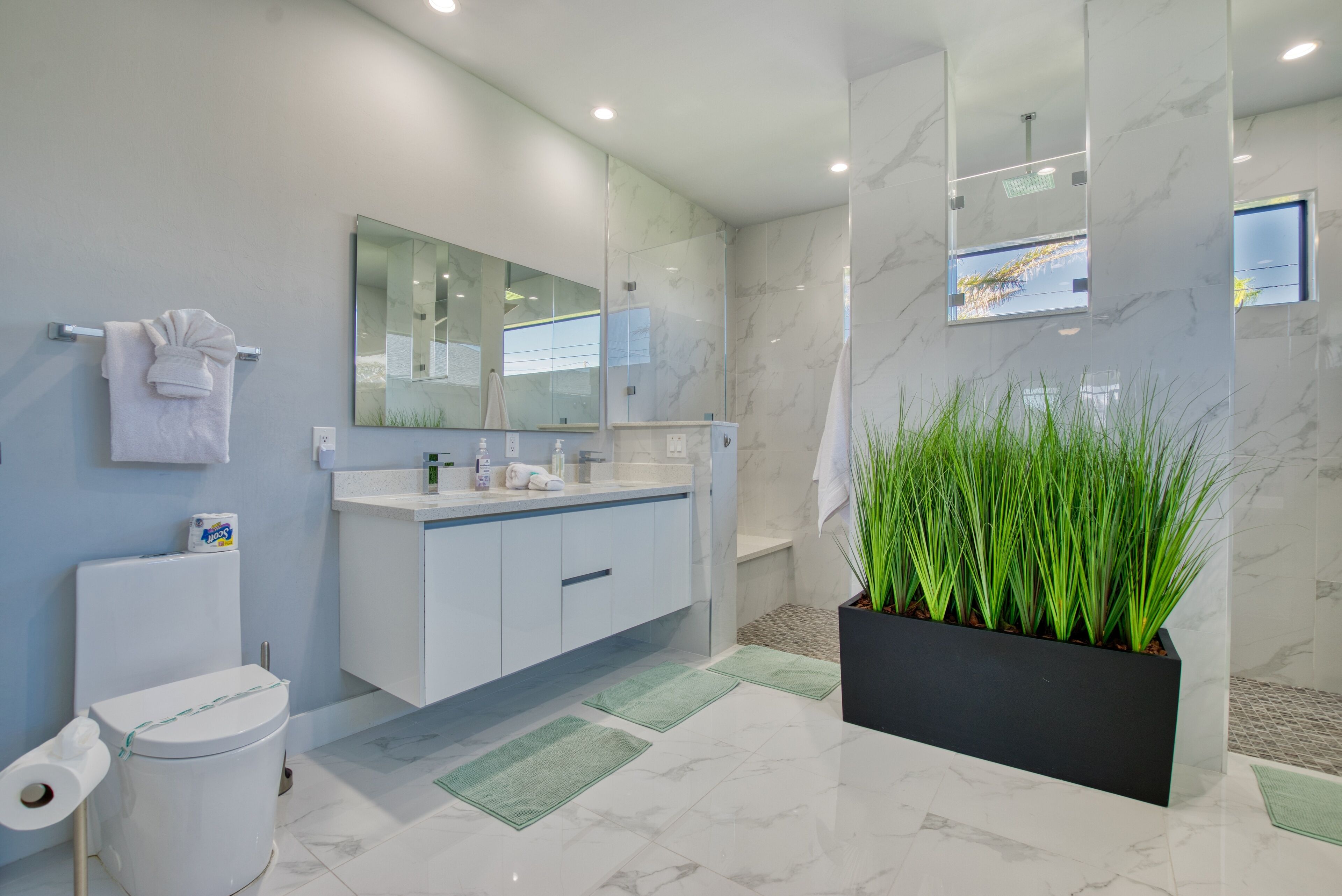 Un baño moderno con inodoro, lavamanos y un espejo grande. Hay una planta en una maceta negra y una toalla colgada en la pared.