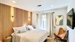 Un dormitorio moderno con cama, pared de paneles de madera, luces de techo, ventana con cortinas, televisor montado y sillón de cuero marrón.