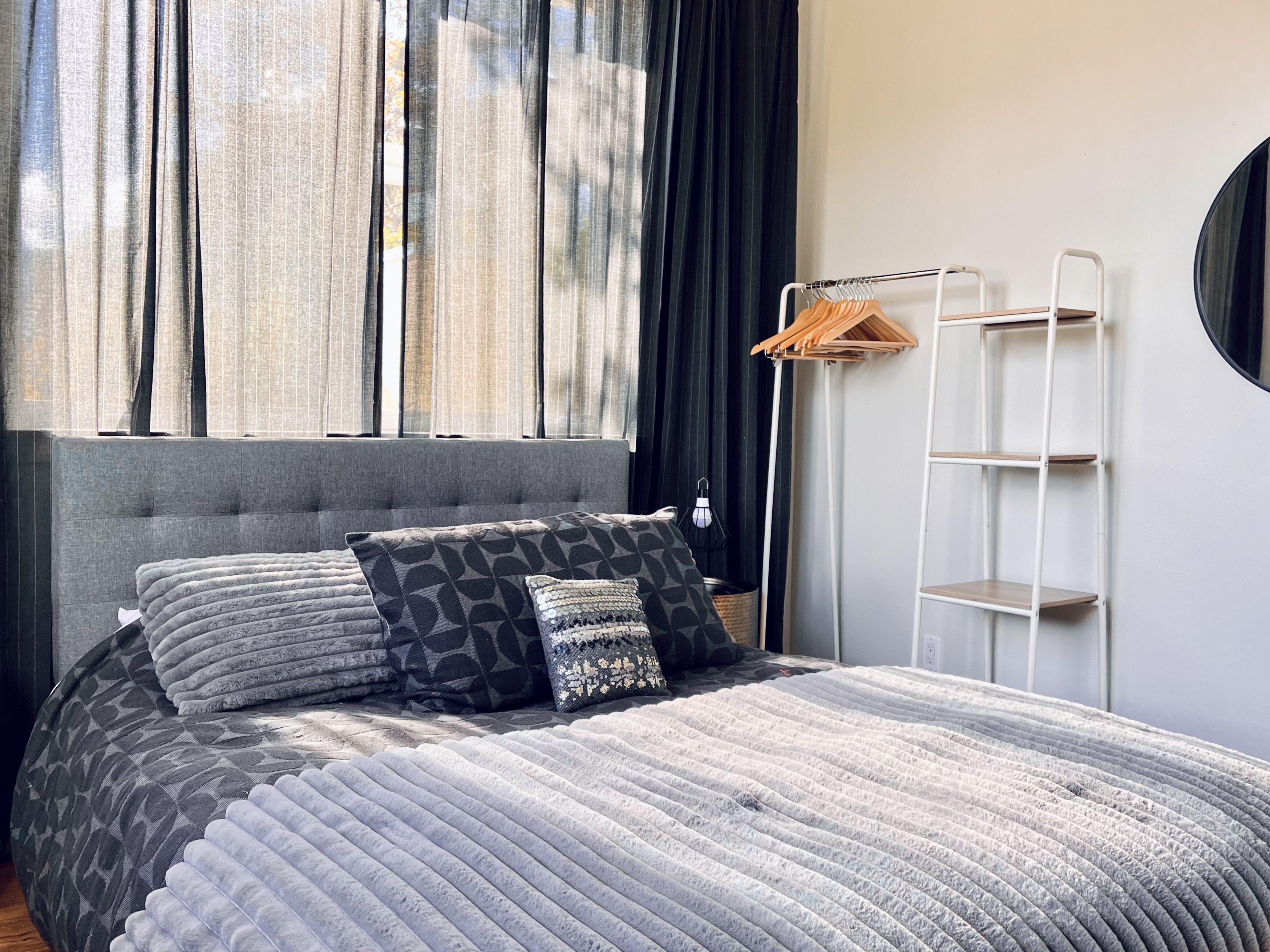 A modern bedroom with a grey upholstered headboard, a grey quilted bedspread, and patterned pillows. A white metal shelf unit is mounted on the wall beside the bed.