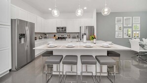 A modern kitchen with stainless steel appliances, a central island with bar stools, pendant lighting, and a dining area with a gray wall and framed pictures. - Vacation Paradise Home, Private Pool, Game Room (Kissimmee)