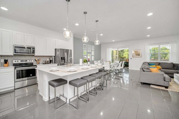 Uma cozinha moderna com armários brancos, eletrodomésticos de aço inoxidável, uma ilha central com banquetas e iluminação pendente.