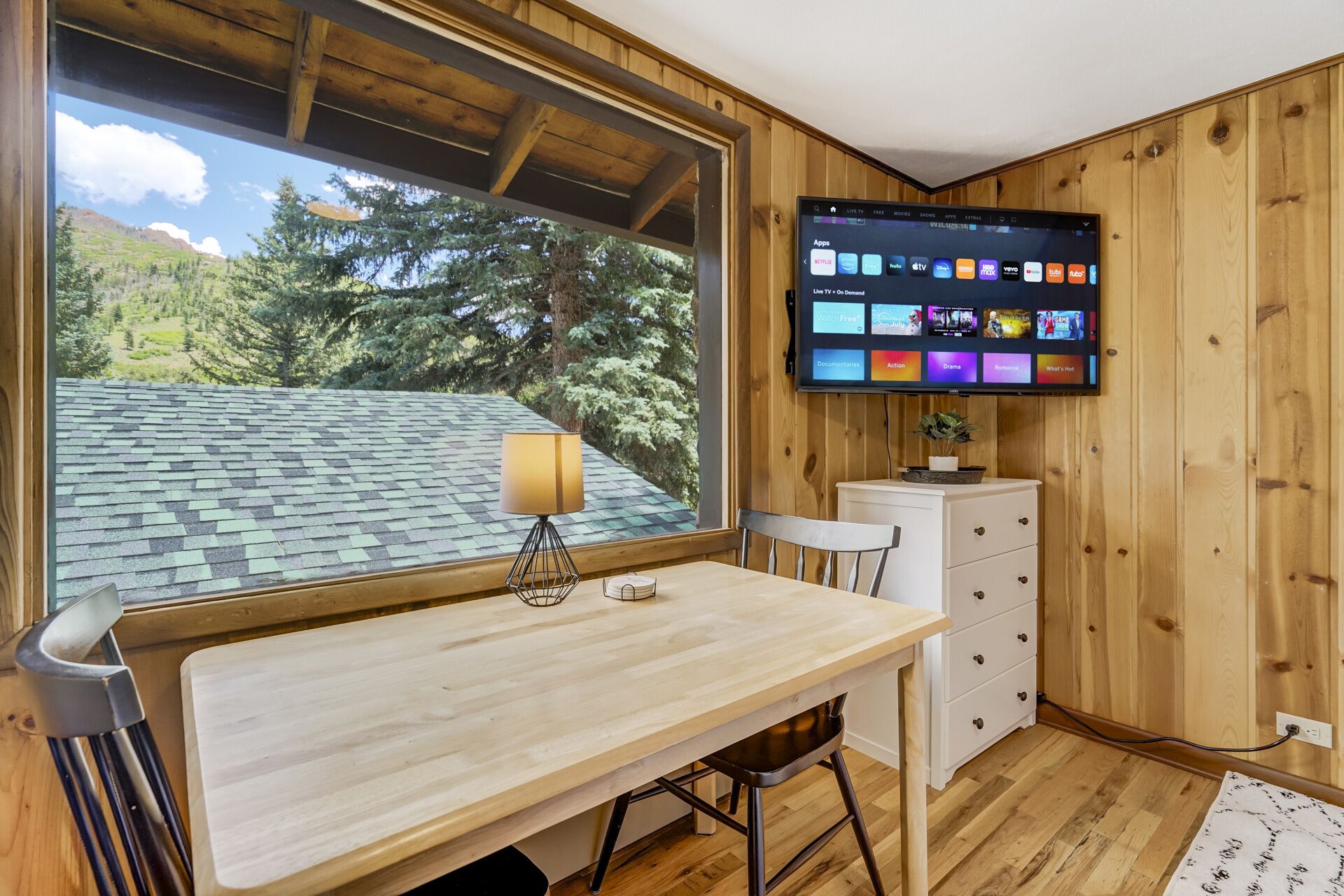 Ein Zimmer mit Holzvertäfelung, einem großen Fenster mit Blick auf Bäume und Berge, einem Schreibtisch mit Lampe, einem Sessel und einem an der Wand montierten Fernseher, der verschiedene Apps anzeigt.