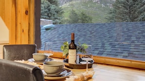 Een eettafel met borden, kommen, een fles wijn en glazen, voor een raam met uitzicht op een bergachtig landschap.