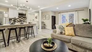 Eine moderne Küche mit einer zentralen Kochinsel, Barhocker, Essbereich und einem Wohnzimmer mit Sofa und Couchtisch.
