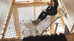 Une personne est assise dans un hamac, dans une pièce dotée d’une grande fenêtre, d’un plafond en bois et offrant une vue sur un paysage enneigé.