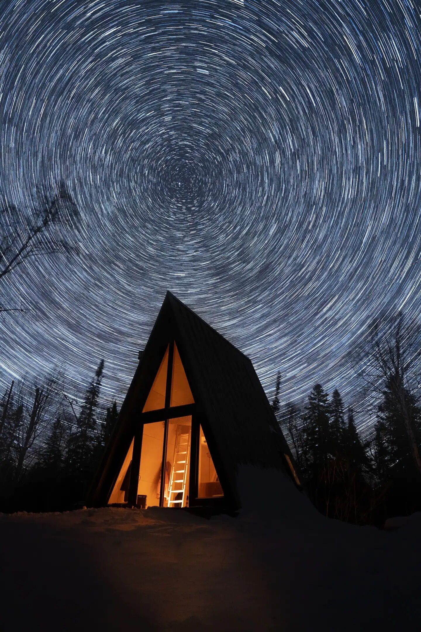 Una cabaña triangular con un ventanal grande, rodeada de nieve, bajo un cielo estrellado.