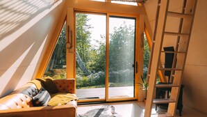 Un intérieur chaleureux et moderne, avec un plafond en pente, une échelle en bois, un canapé agrémenté de coussins et une grande fenêtre donnant sur un paysage verdoyant.
