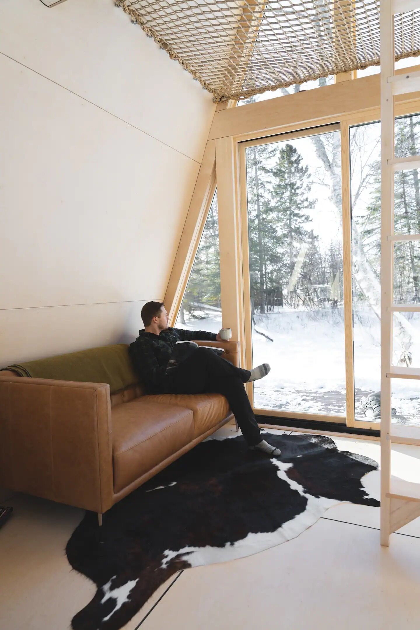 A person sitting on a brown leather couch in a room with a large window, a cowhide rug, and a wooden ceiling structure.
