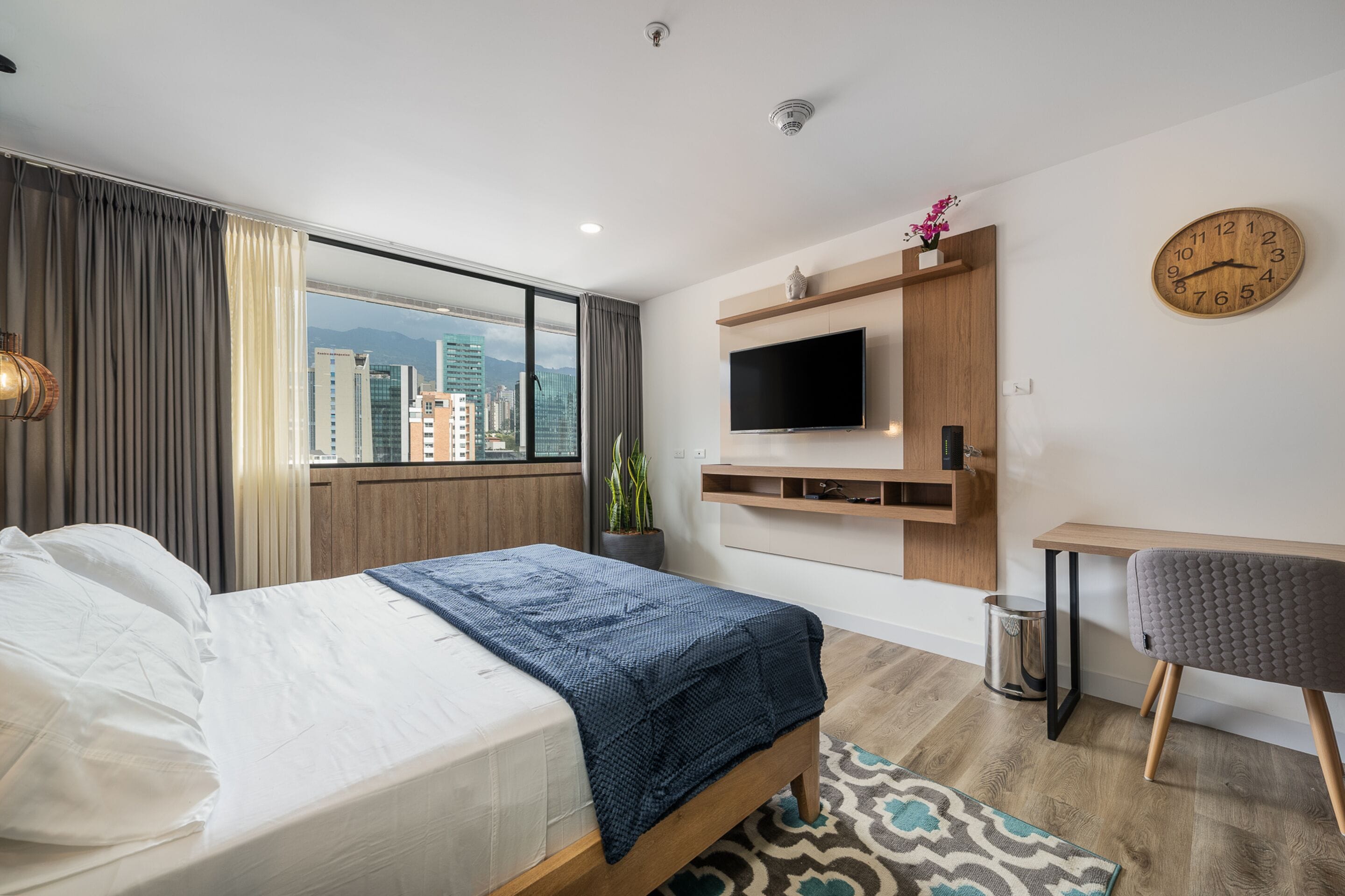 A modern hotel room with a large bed, a TV mounted on the wall, a small desk, and a view of the city.