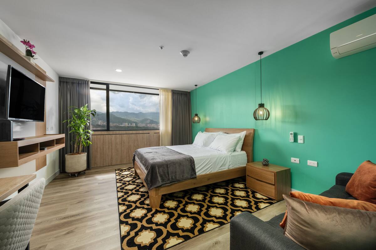 Una habitación de hotel moderna con una cama grande, televisión, escritorio, silla, planta en maceta y vistas a la ciudad.
