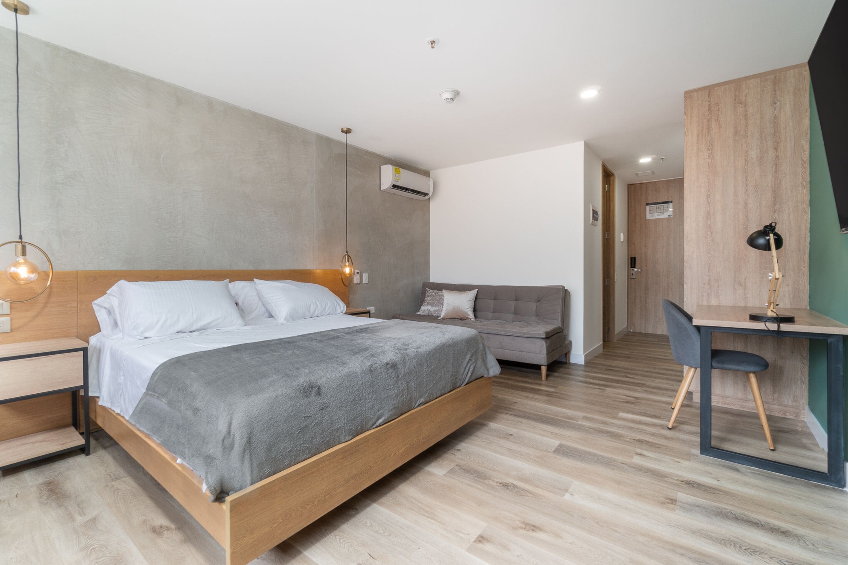 A modern bedroom with a wooden bed, a grey sofa, a desk with a chair, and a hanging light fixture.