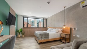 A modern hotel room with a large bed, a desk, a chair, a TV mounted on the wall, and a view of the cityscape through the window.