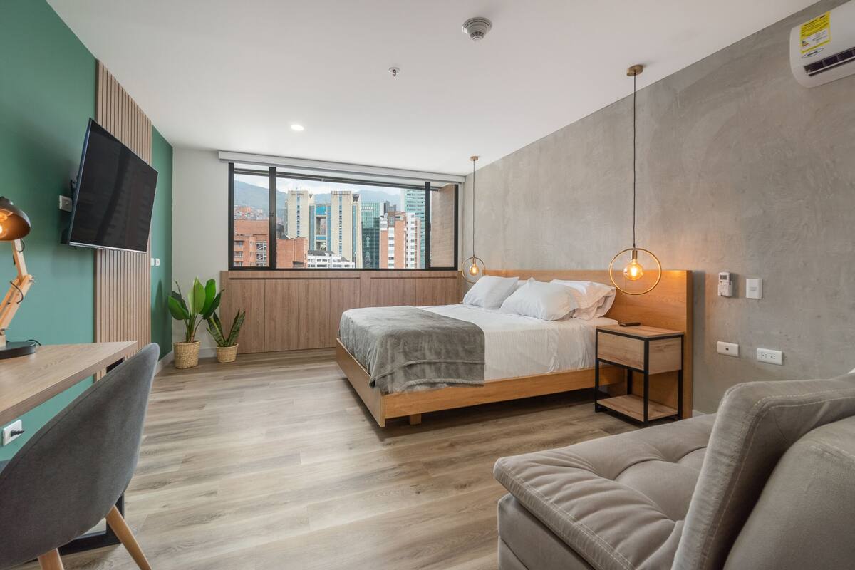 Una habitación de hotel moderna con una cama grande, un escritorio, una silla, un televisor montado en la pared y vistas de la ciudad a través del ventanal.