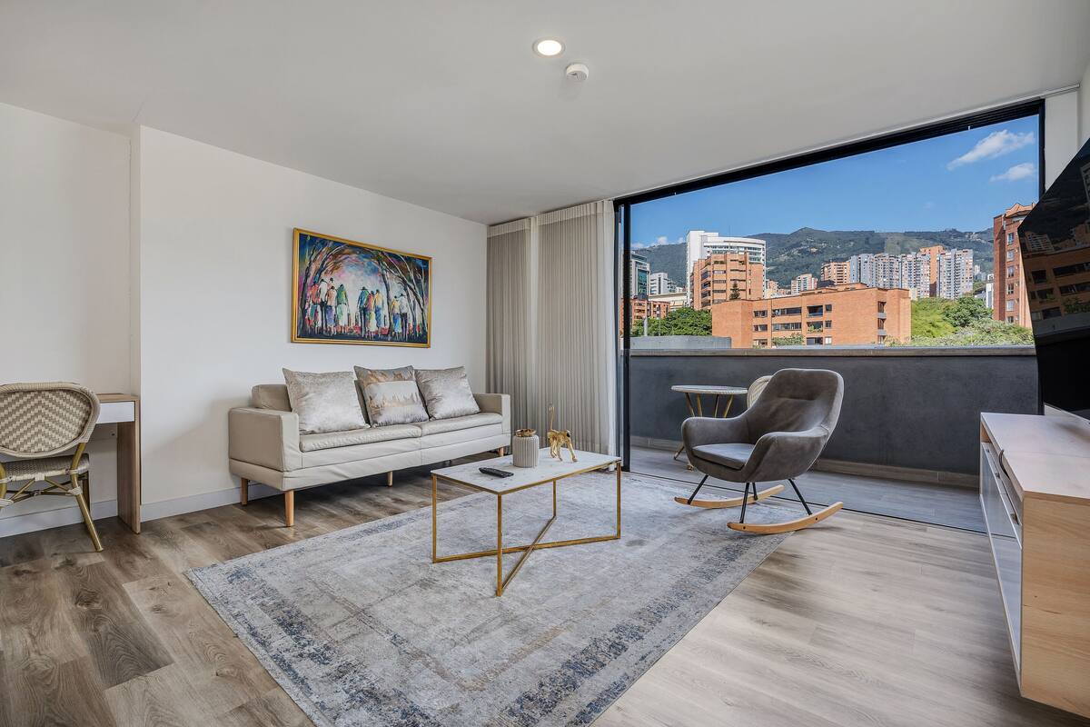 Una sala de estar moderna con un sofá, una mesa de centro y un sillón mecedora. Hay un cuadro en la pared y una vista de edificios a través de un ventanal grande.