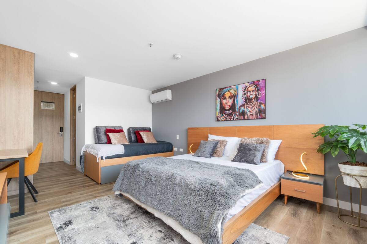 Un dormitorio moderno con una cama de madera, un escritorio y una silla. Hay un cuadro en la pared y una planta al lado.