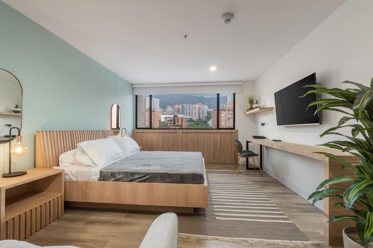 Un dormitorio moderno con cama, mesita de noche y escritorio con silla. Hay una ventana grande con vista a la ciudad.