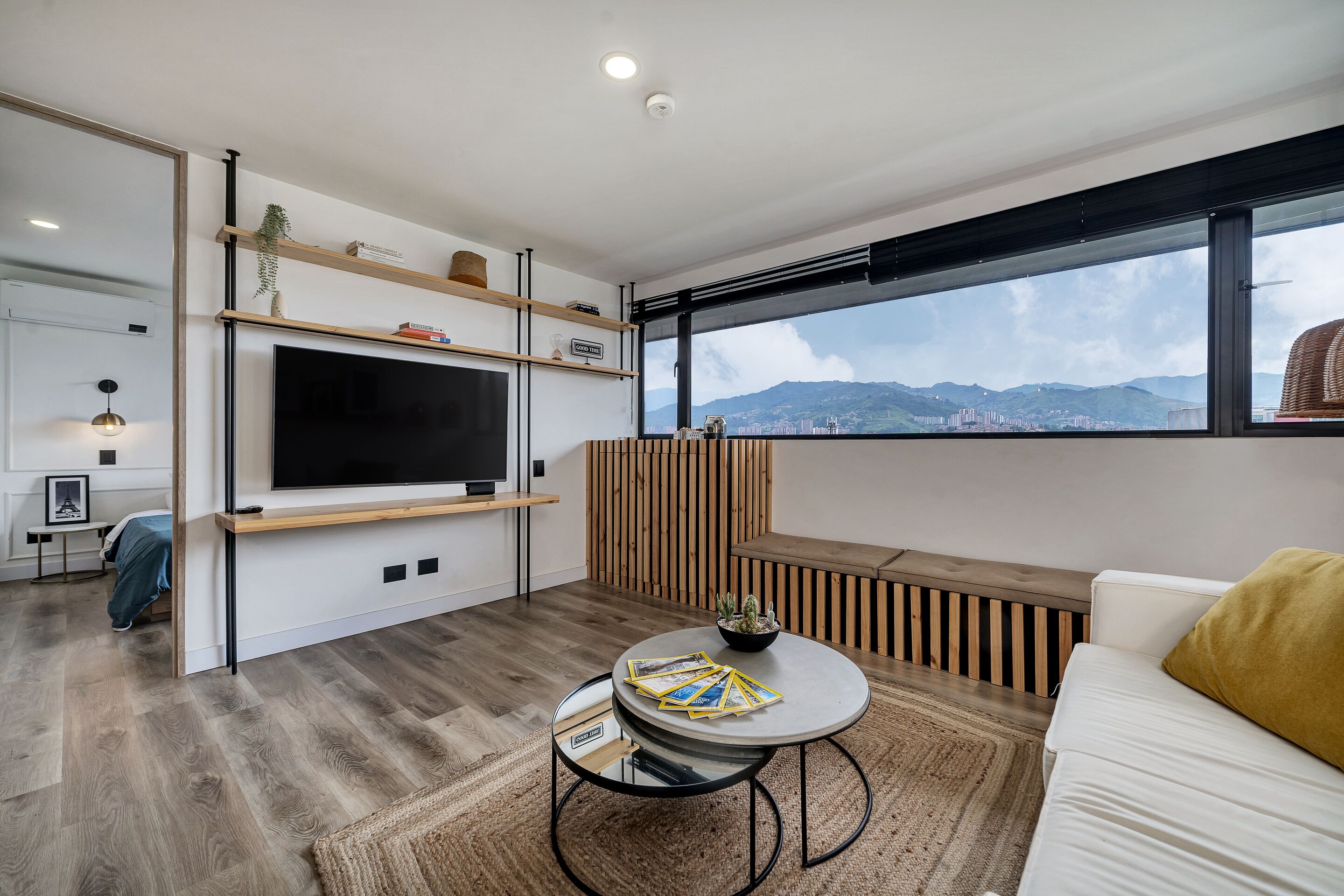 Een moderne woonkamer met een flatscreen televisie, houten planken, een ronde salontafel en een stadsgezicht door grote ramen.