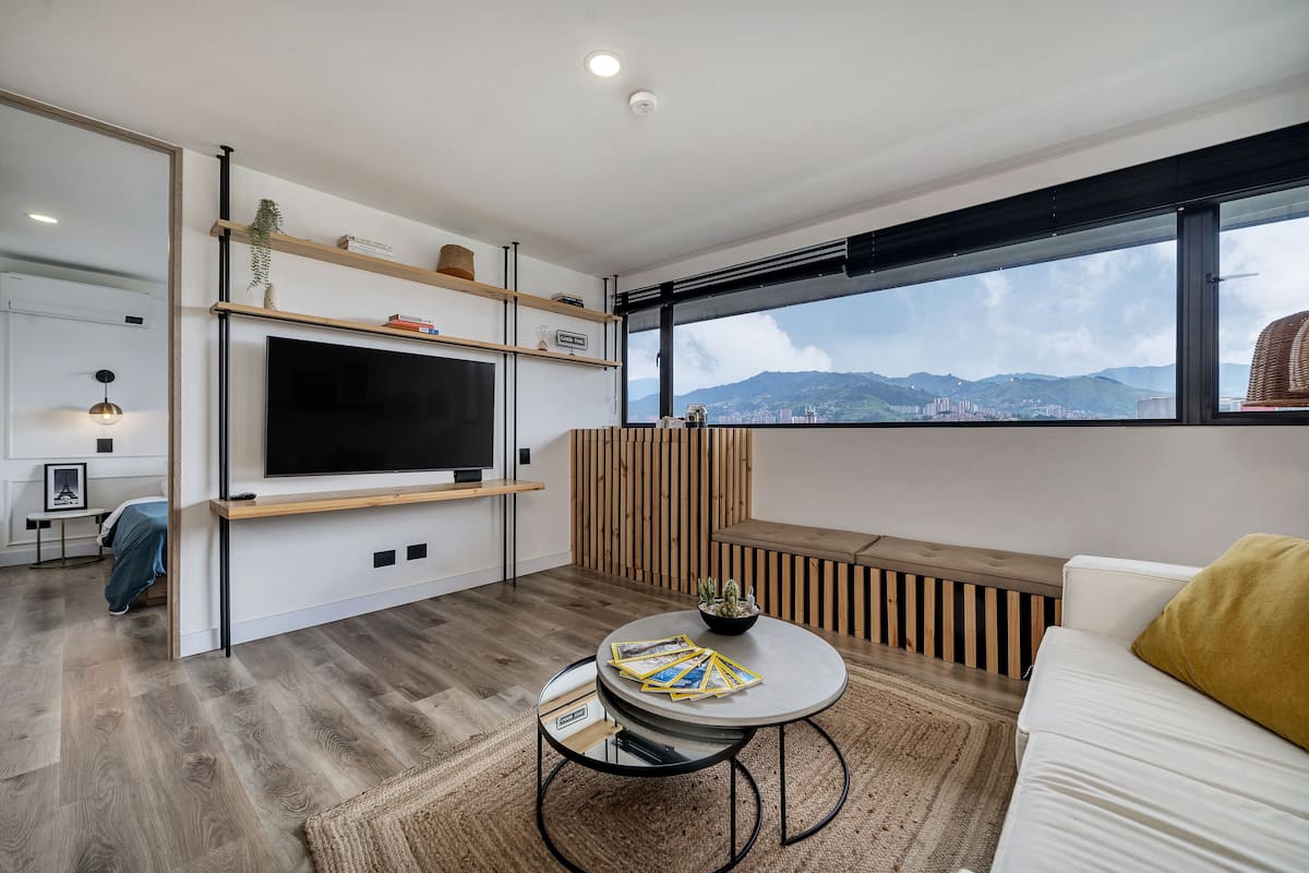 Una sala de estar moderna con una televisión de pantalla plana, estanterías de madera, una mesa redonda de centro y vistas a un paisaje urbano a través de grandes ventanales.