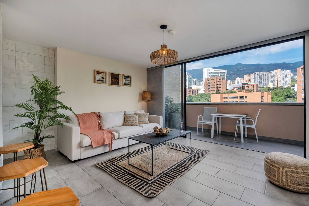 Una sala de estar moderna con un sofá, una mesa de centro y sillas, un ventanal con vistas a la ciudad y una planta en maceta.