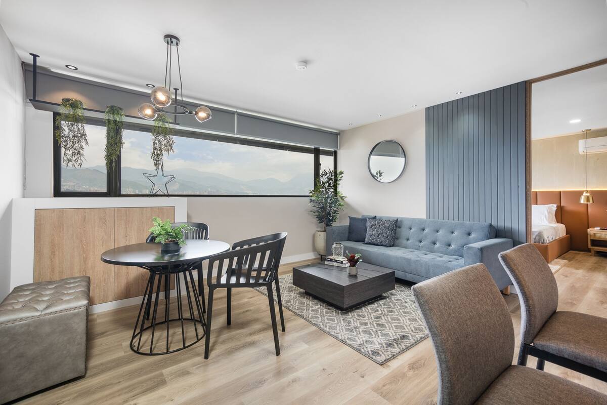 Una sala de estar moderna con un ventanal grande, un espejo redondo, un sofá azul, una mesa de centro y una zona de comedor con mesa y sillas.