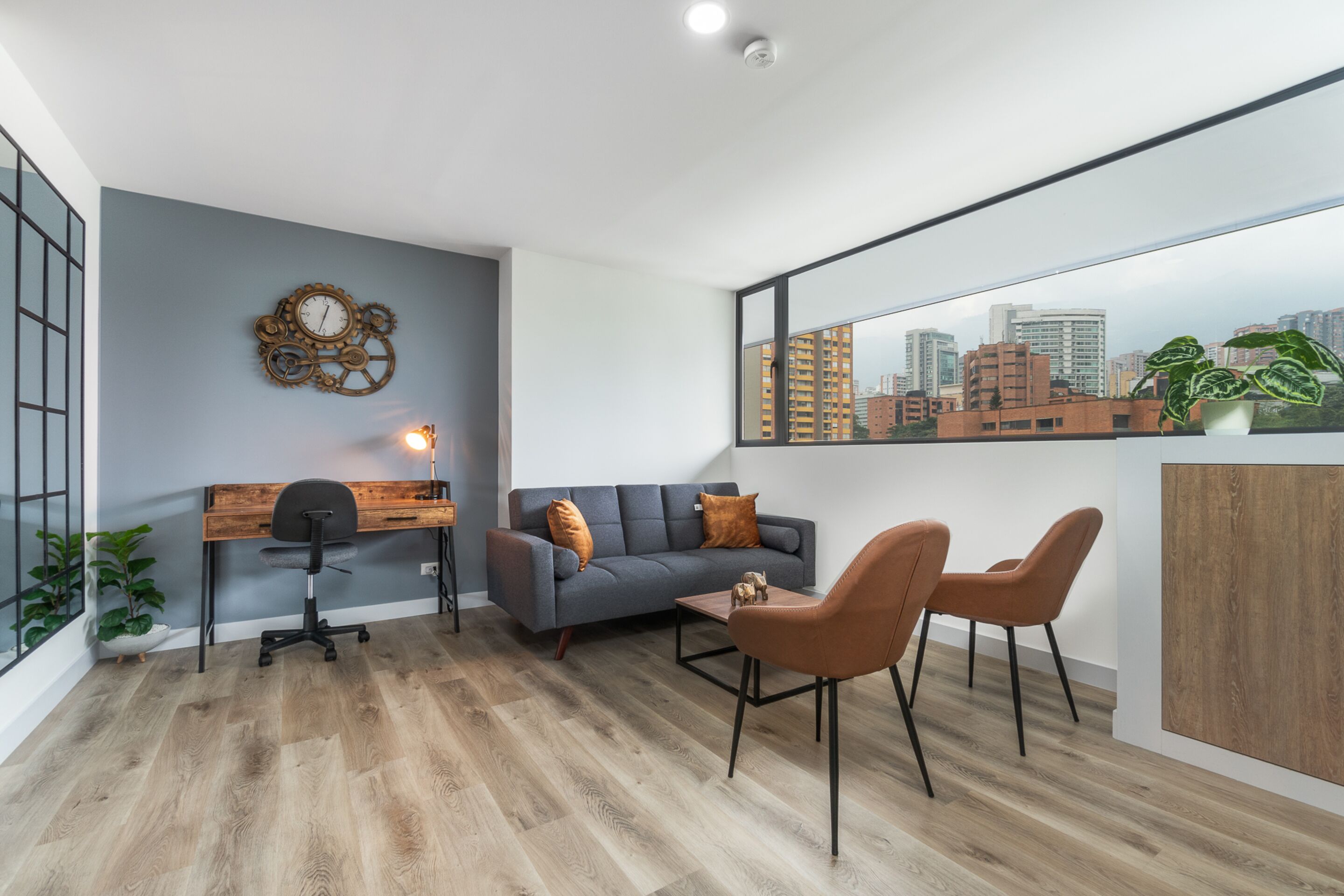 A modern living room with a grey sofa, two brown chairs, a wooden desk, and a city view through a large window.