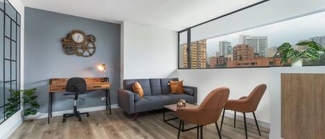 A modern living room with a grey sofa, two brown chairs, a wooden desk, and a city view through a large window.